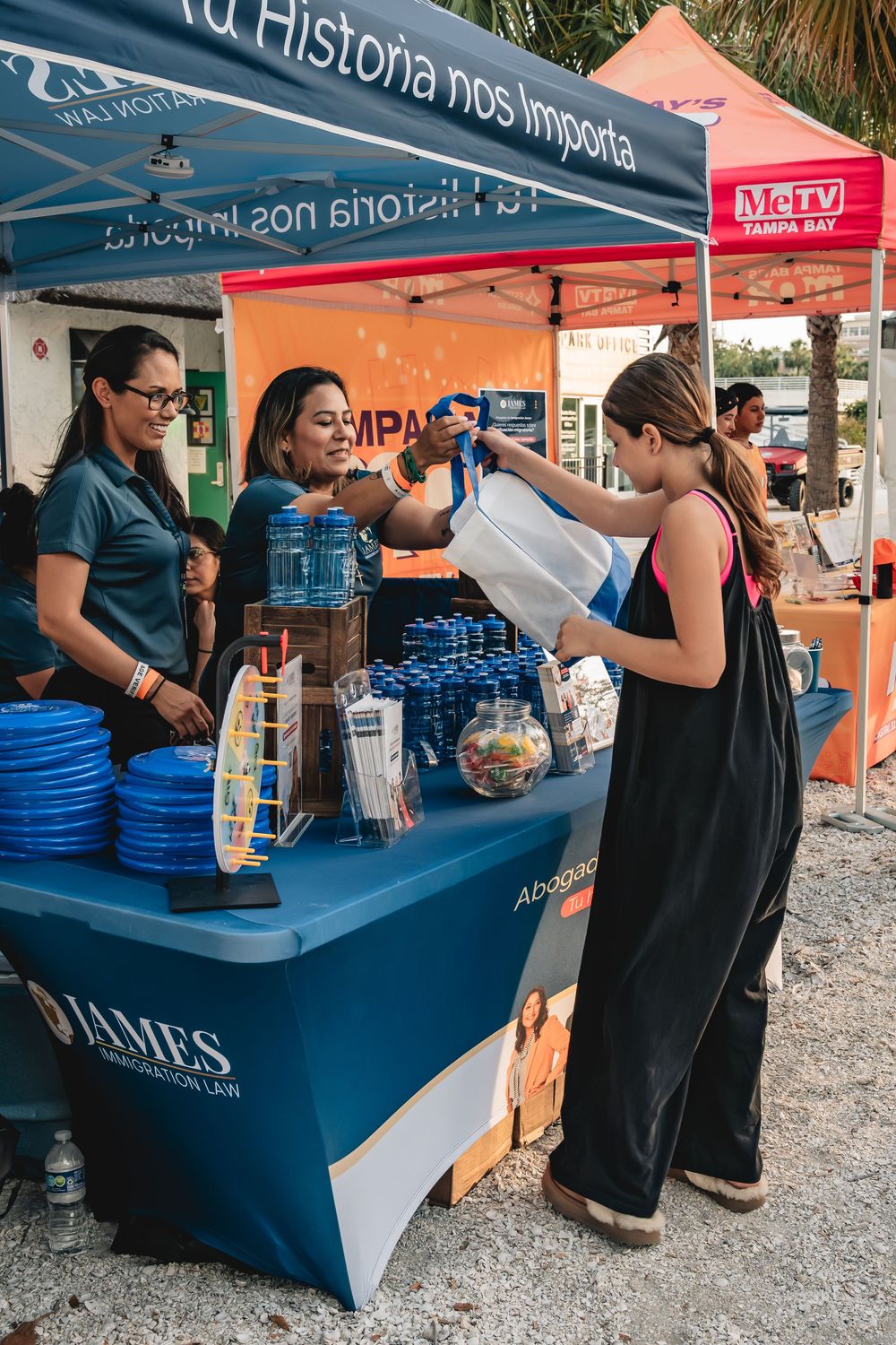 Women serving water at a James Immigration Law community event booth in Tampa Bay under a blue canopy tent.