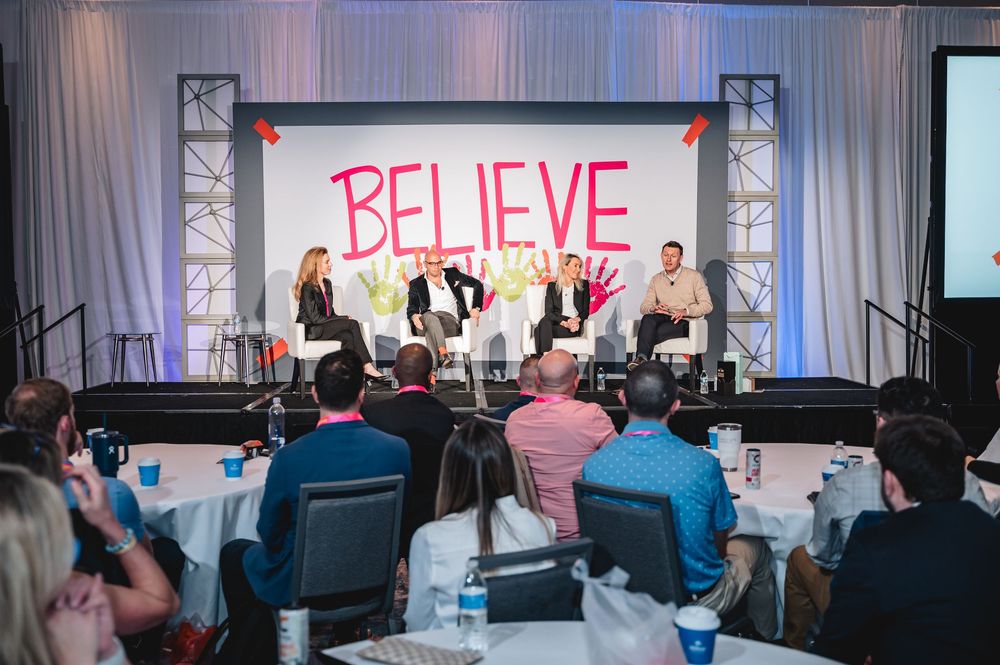 Panel discussion on stage at a Believe conference with five speakers seated before an audience at round tables.