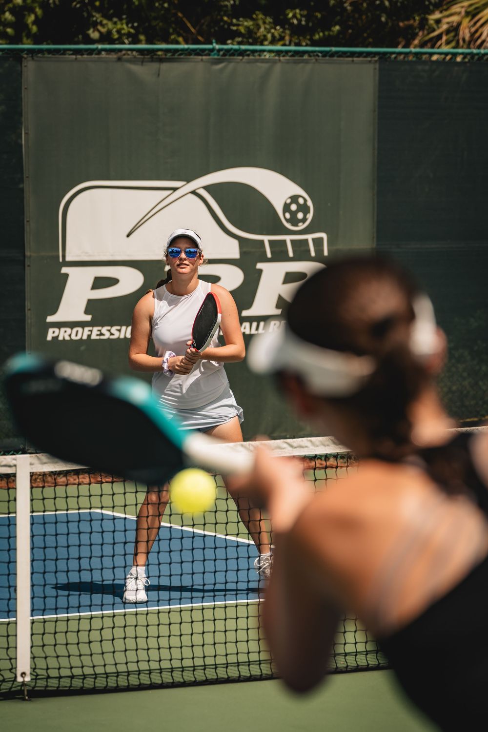 Two women playing pickleball on an outdoor court with a PPA logo banner in the background.