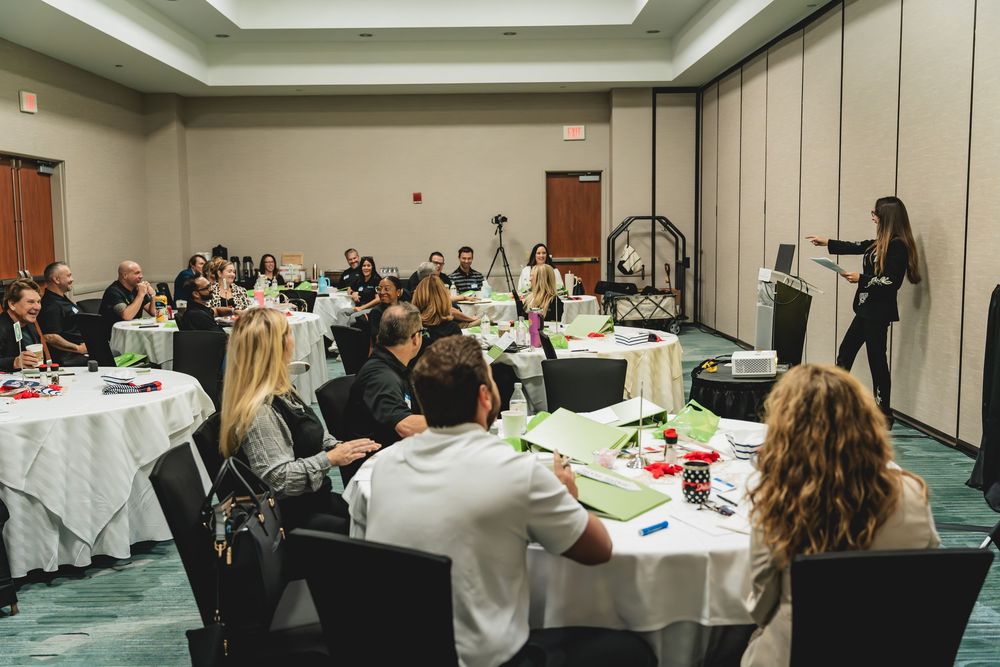 Professional seminar with diverse attendees seated at round tables listening to a female presenter in a conference room.