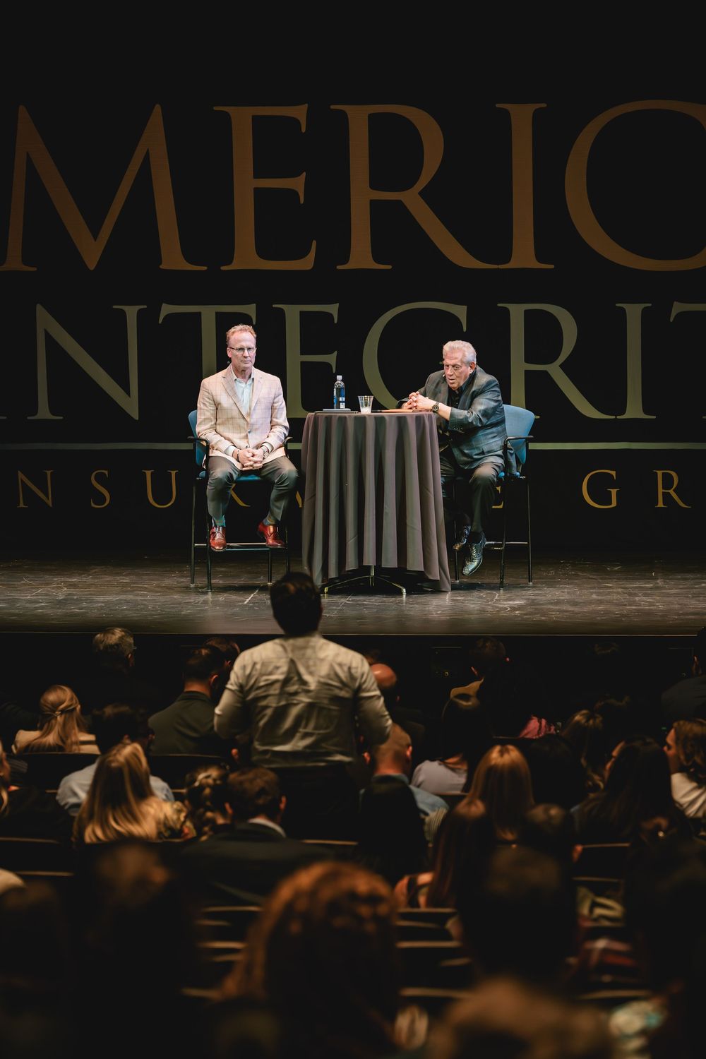 Two speakers on stage at American Integrity Insurance Group event, seated at table before large audience in theater.