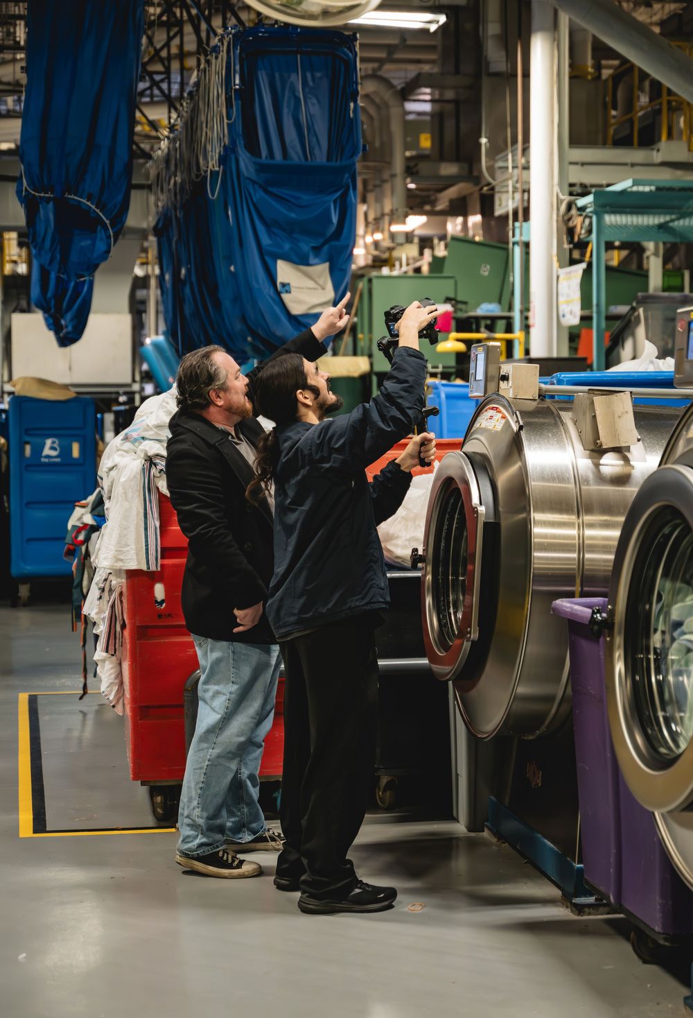 Two workers inspecting industrial laundry equipment in a large commercial laundry facility.