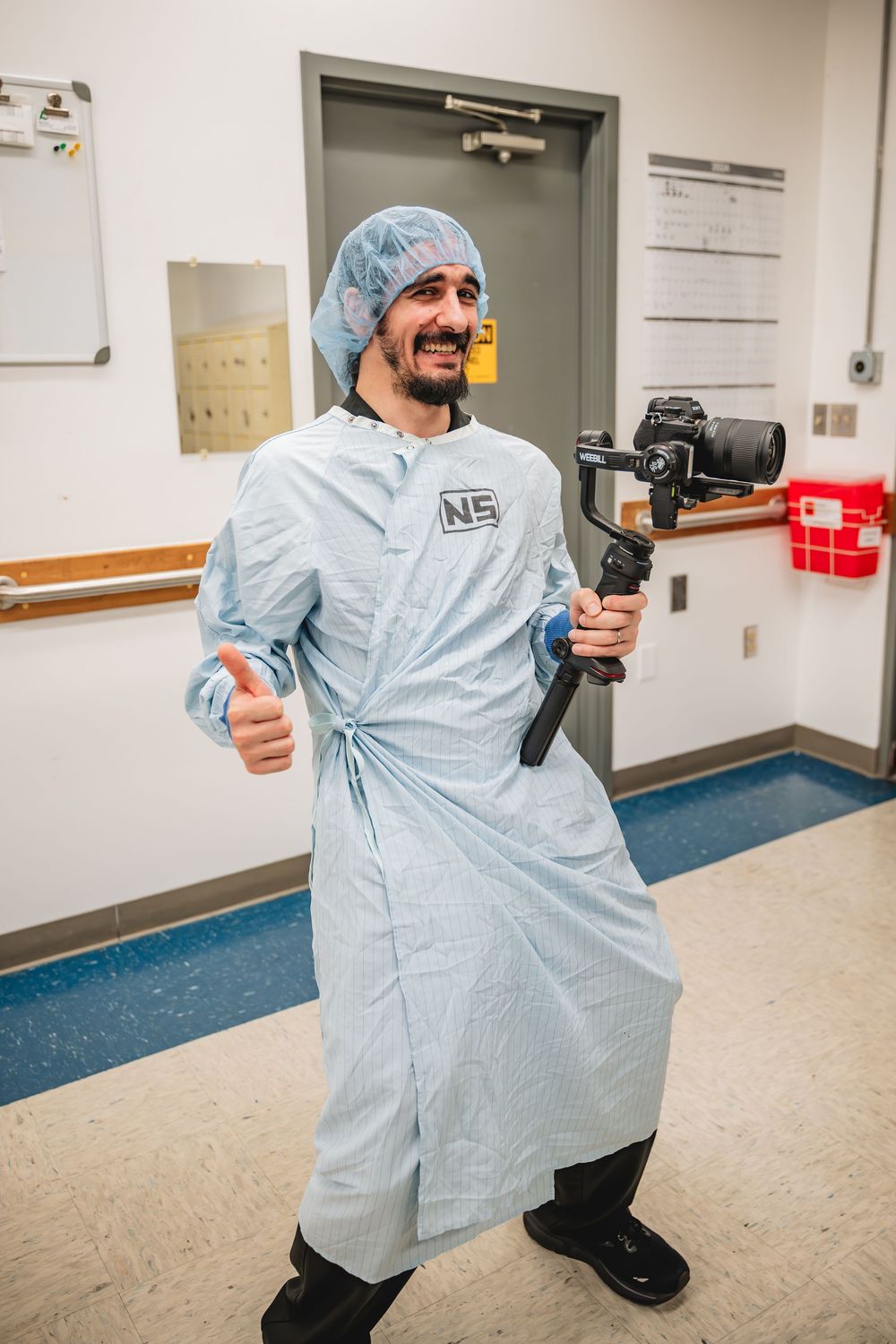 Man in surgical gown and hair net holds a camera gimbal stabilizer, giving thumbs up in a hospital hallway.