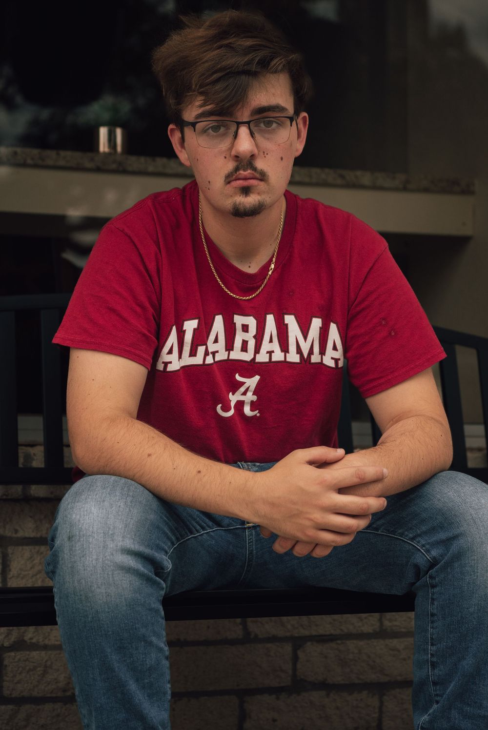 Young man in red Alabama t-shirt and jeans sits outdoors, wearing glasses and a gold chain.