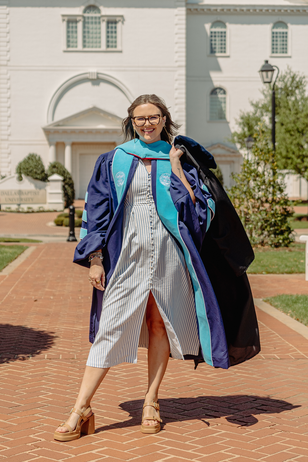 Woman in doctoral graduation regalia walks on brick path outside white university building.