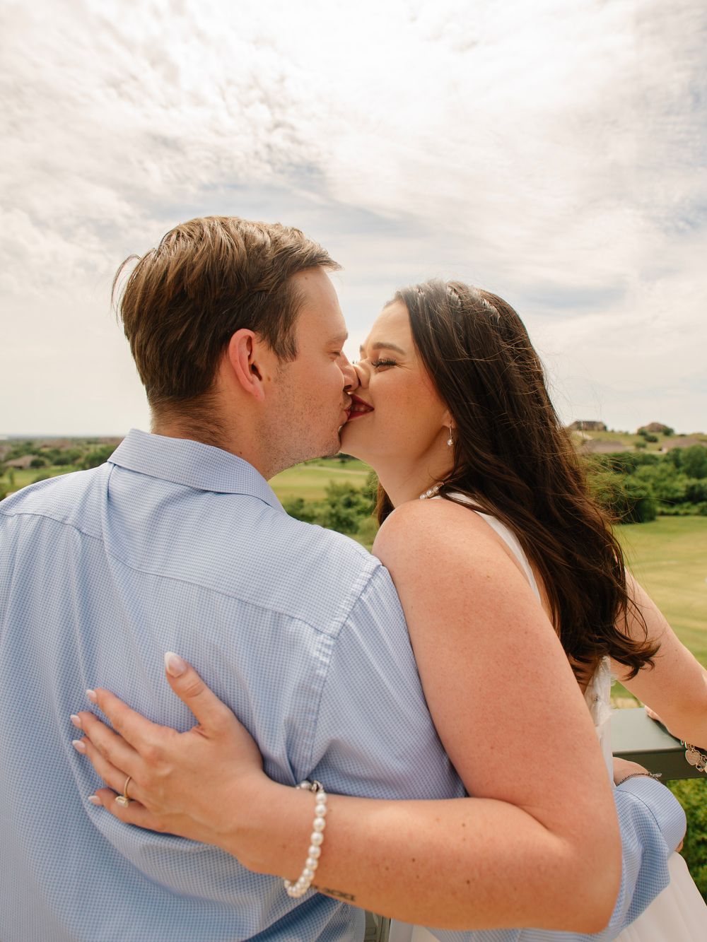 Couple sharing a romantic kiss outdoors with lush green landscape and cloudy sky in the background.
