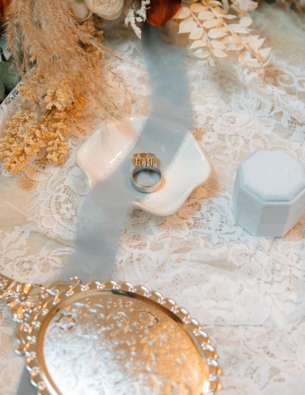 Wedding ring resting on a white ceramic holder atop lace fabric, surrounded by dried florals and a silver tray.