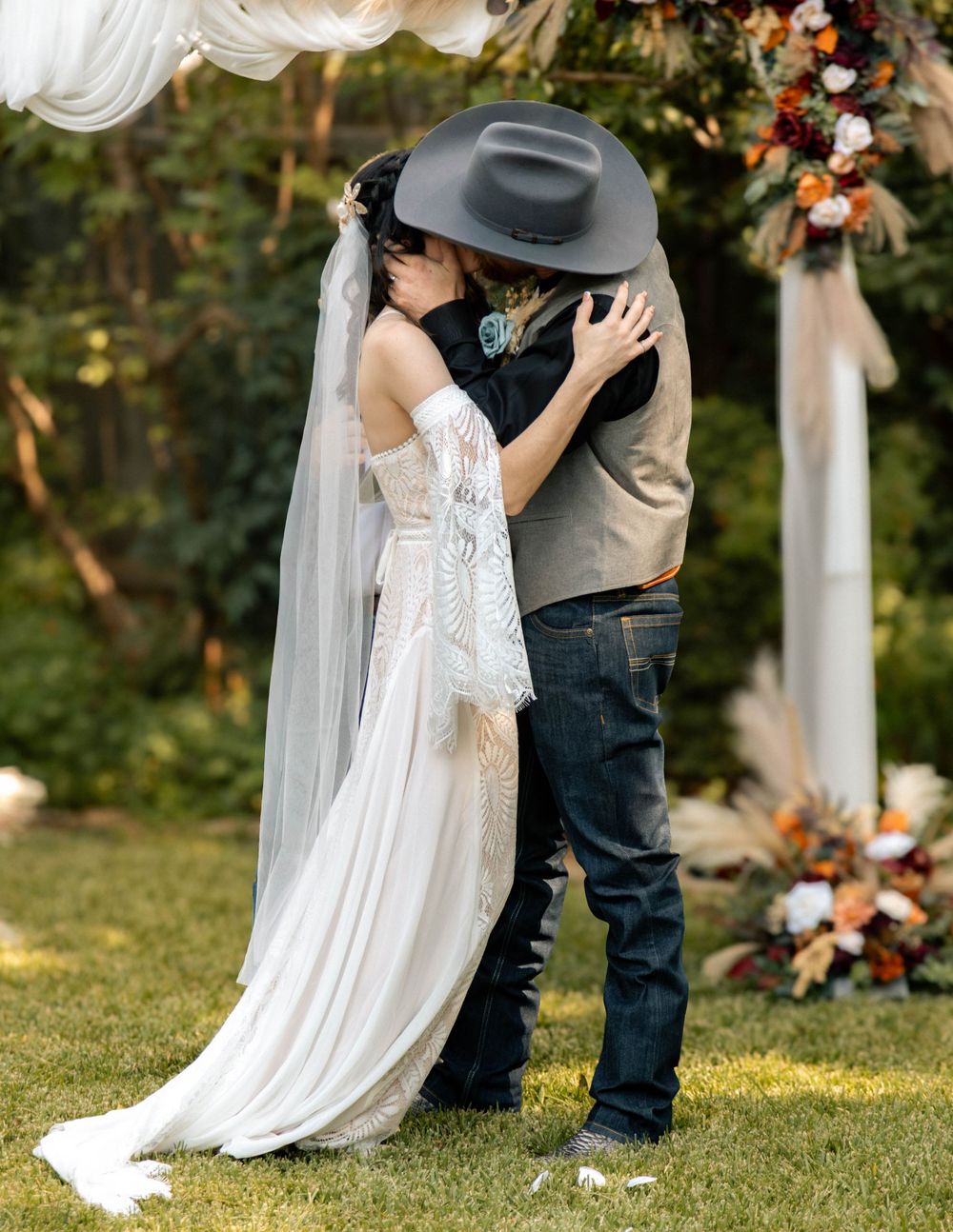 Bride and groom share first kiss at outdoor western wedding, bride in lace gown with veil, groom in cowboy hat and jeans.