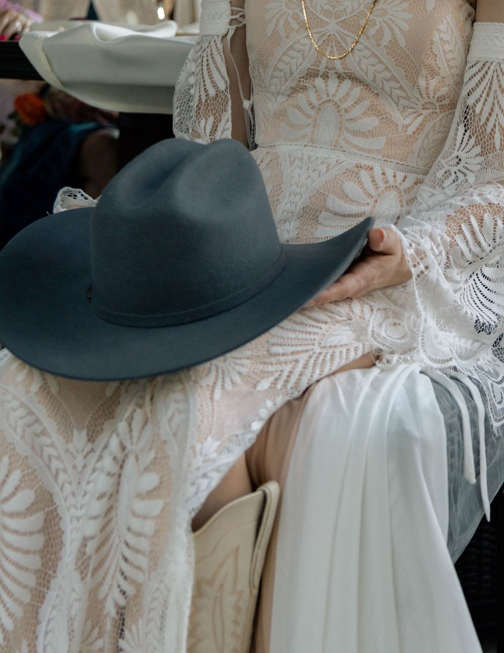 A bride in a lace wedding gown holds a wide-brim gray felt hat while wearing a delicate gold necklace.