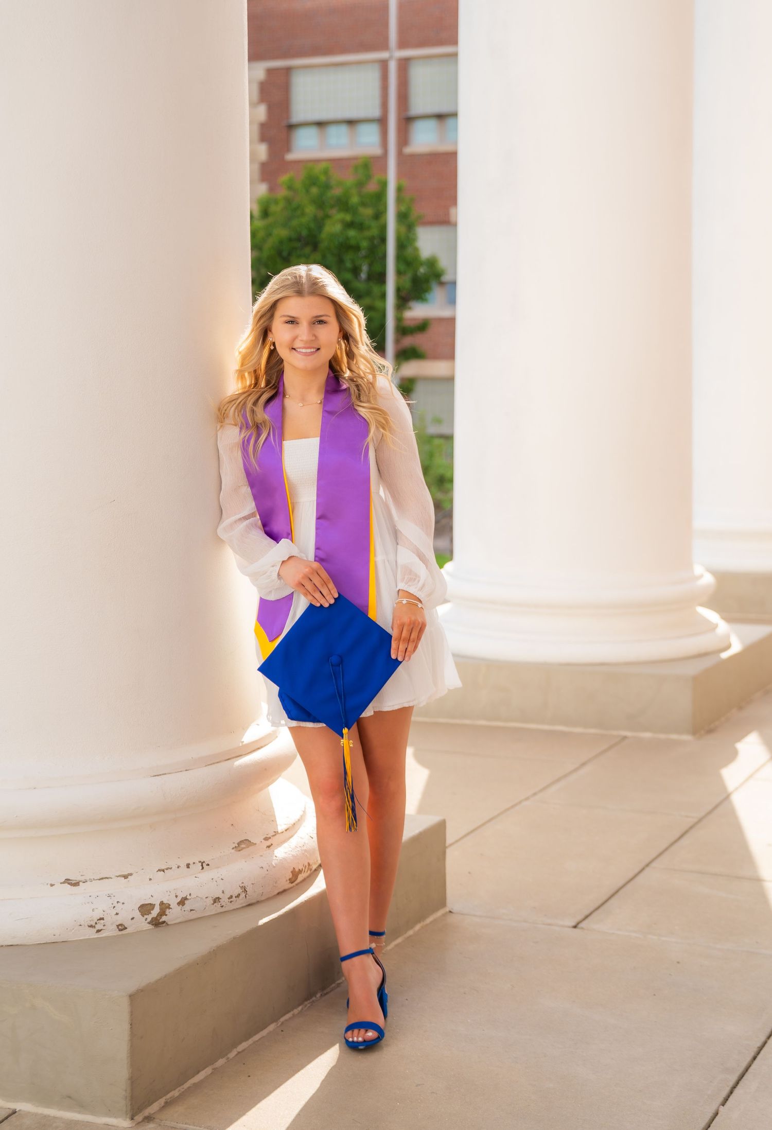 Smiling female graduate in white dress with purple and gold stole holding blue cap between white columns.