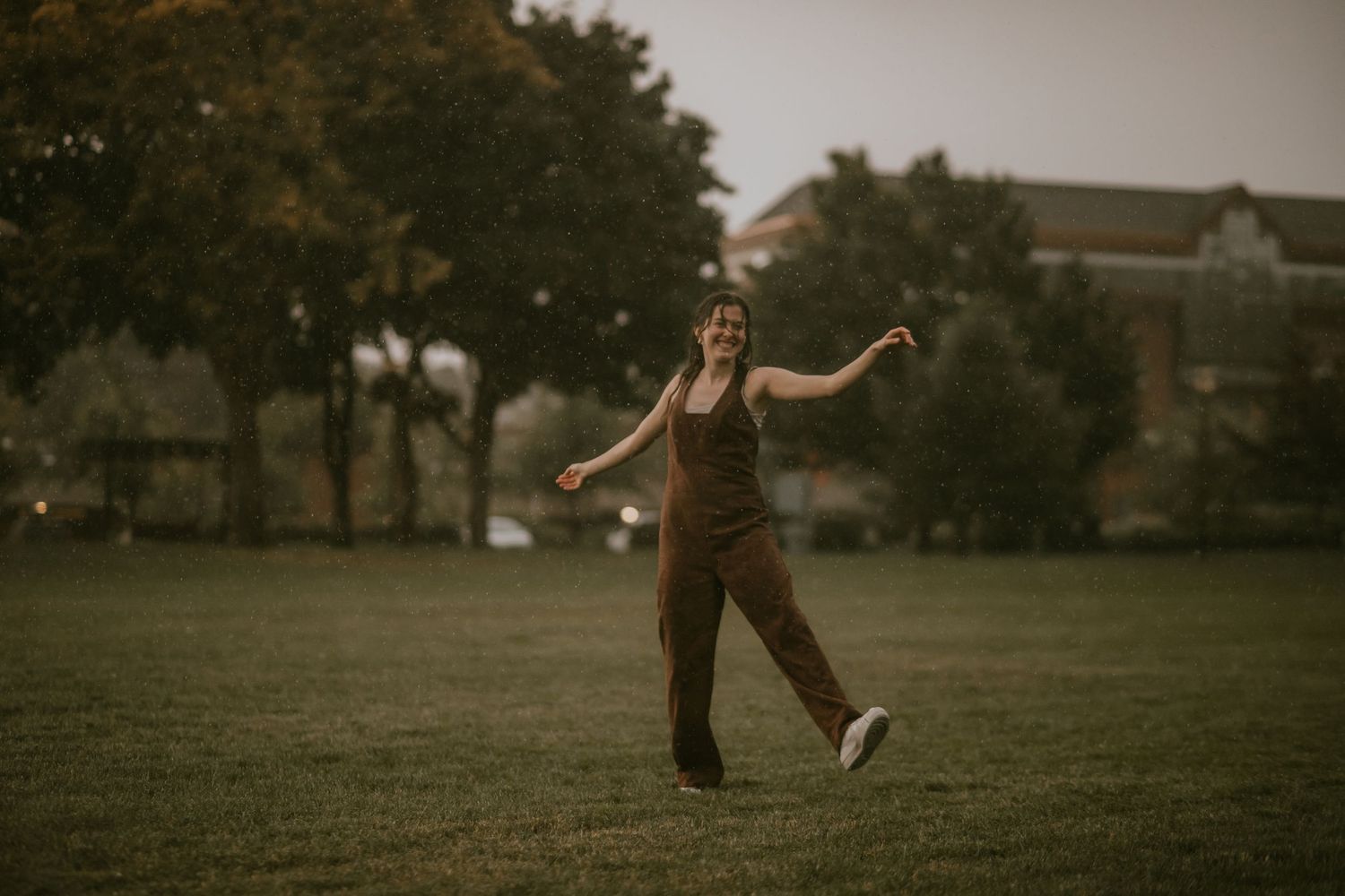 Person in brown overalls dancing joyfully in a green park at dusk with trees and a building in the background.
