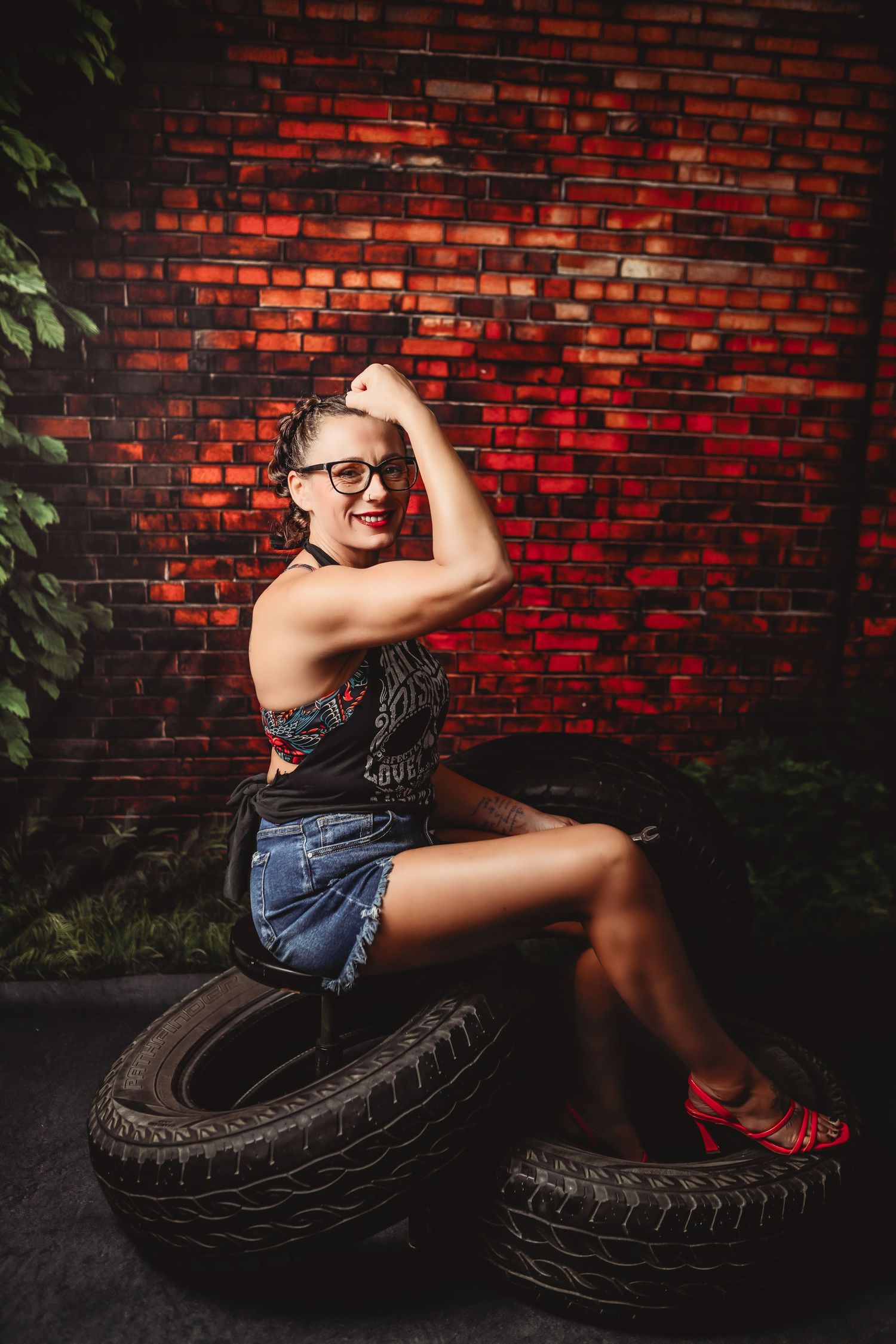 Redhead woman in plaid skirt, black blazer, and red heels sitting on vintage desk against brick wall.