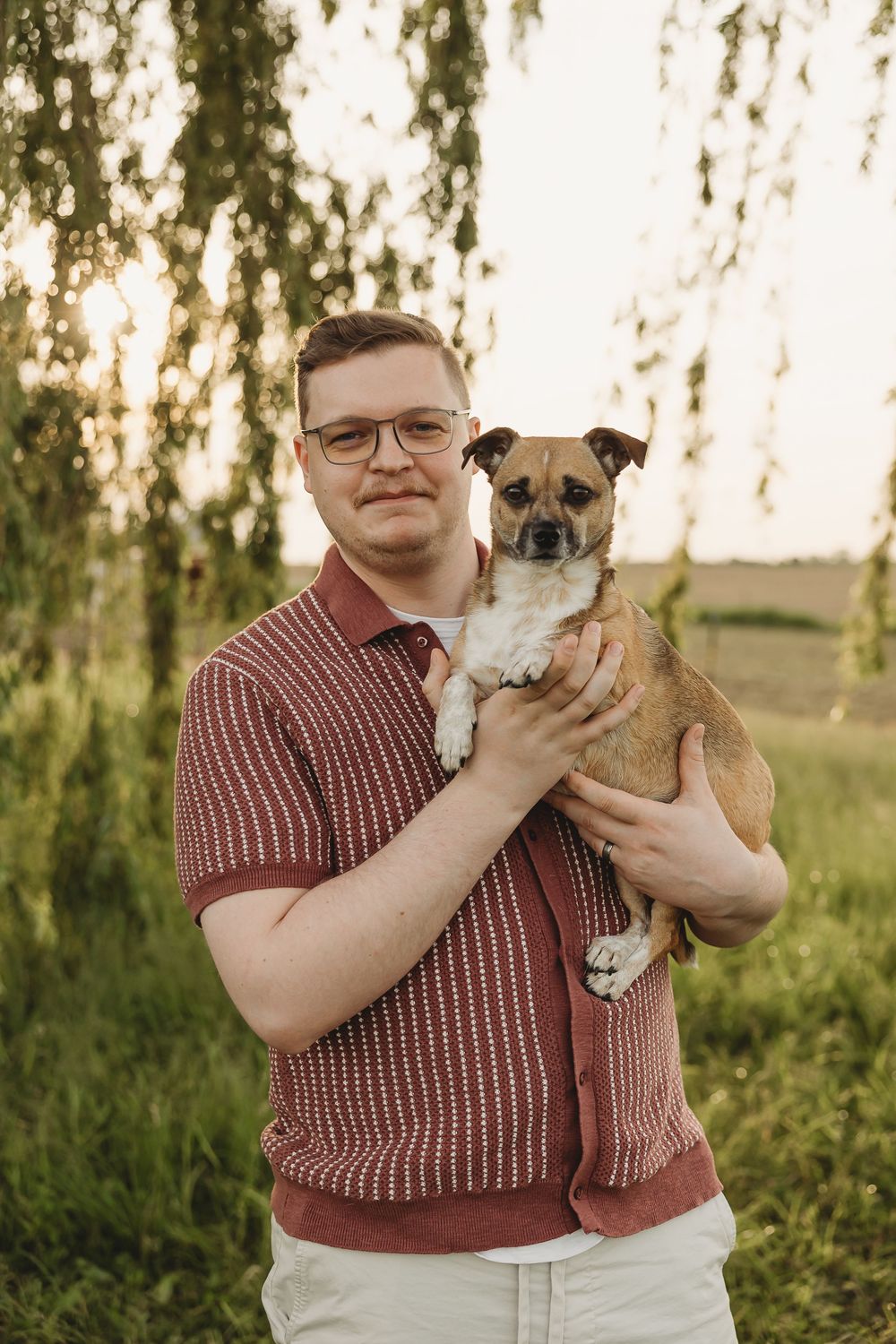 Man in red polo shirt holding a small brown dog outdoors with green trees in golden sunset light.