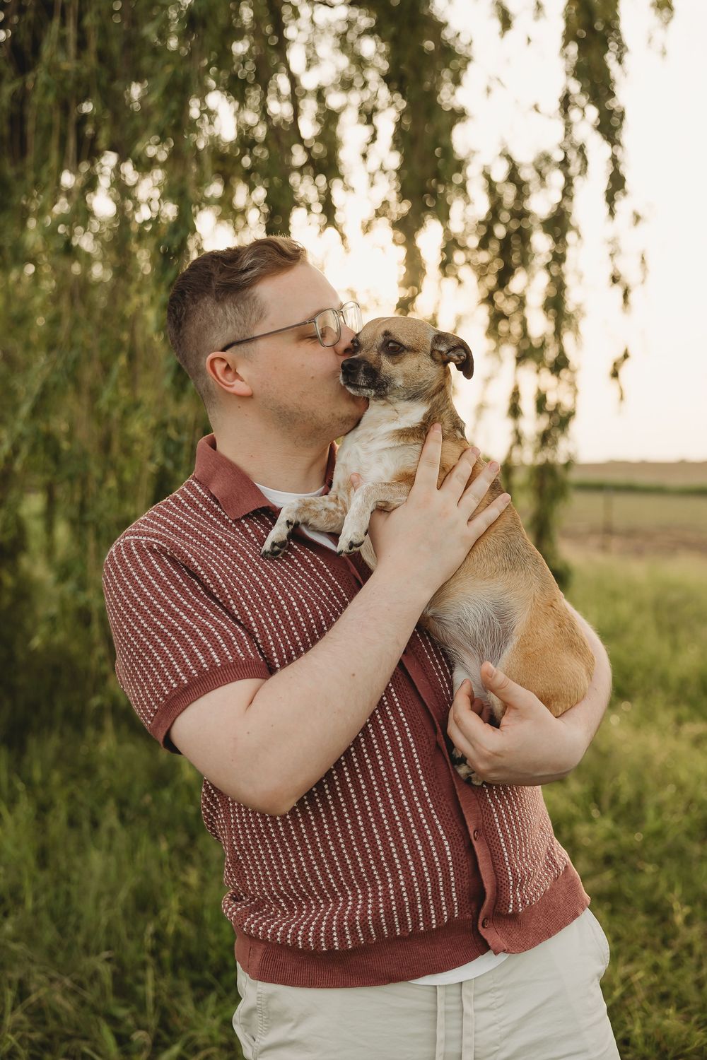 Man in red polo shirt kissing small brown dog outdoors near weeping willow tree at sunset.
