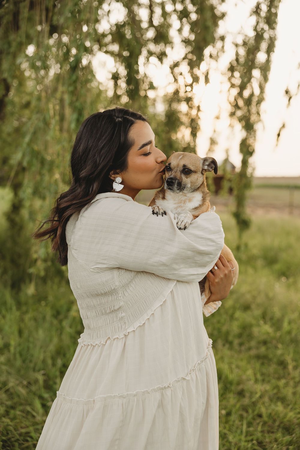 Woman in white dress kissing small chihuahua dog outdoors at sunset near green weeping willow trees.