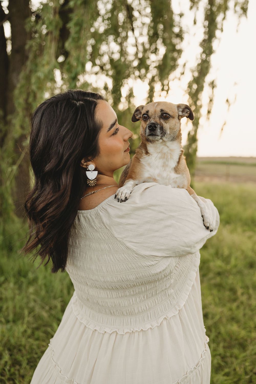 Woman in white off-shoulder dress holds small tan dog near weeping willow trees at sunset.