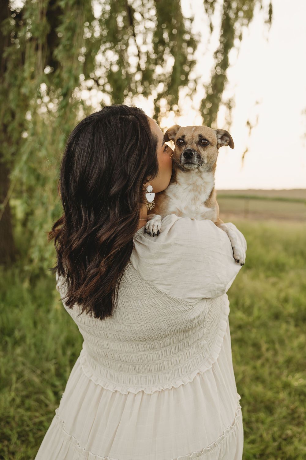 Woman in white dress holding a small dog near a willow tree at sunset.