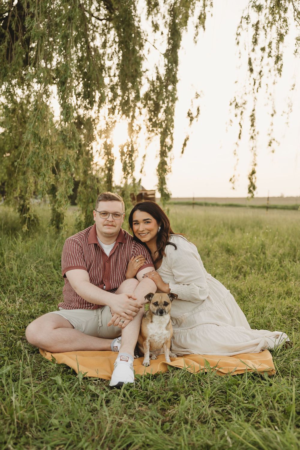 Couple sitting on blanket under willow tree at sunset with small Chihuahua dog in green field.