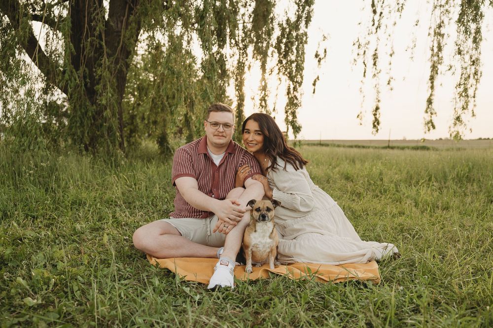 Couple sitting on blanket with small dog under willow tree in green field at sunset.