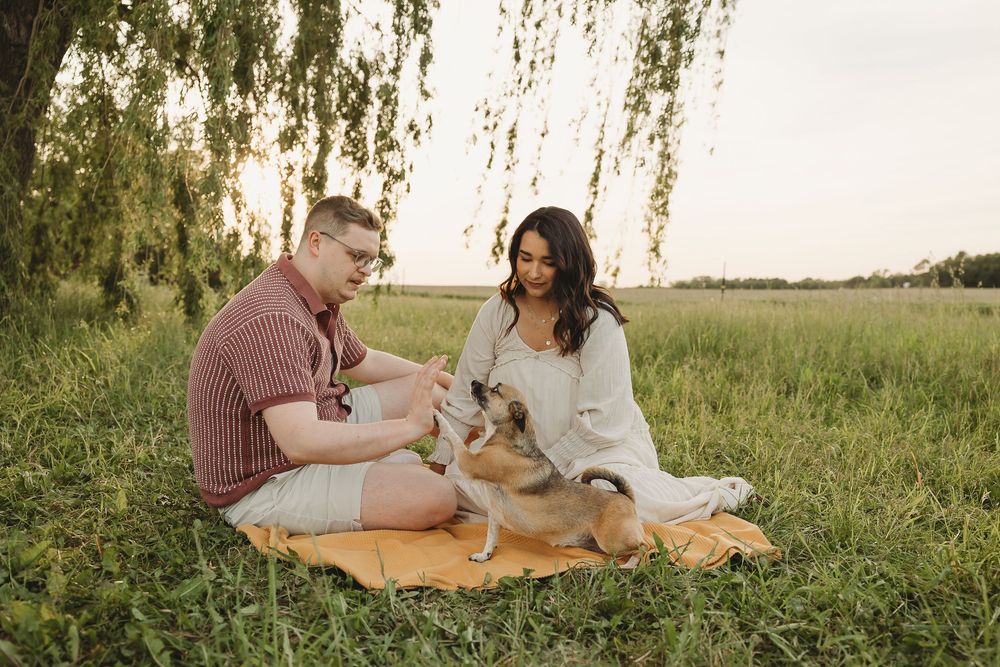 Couple sitting on blanket in green field with their dog at golden hour sunset.