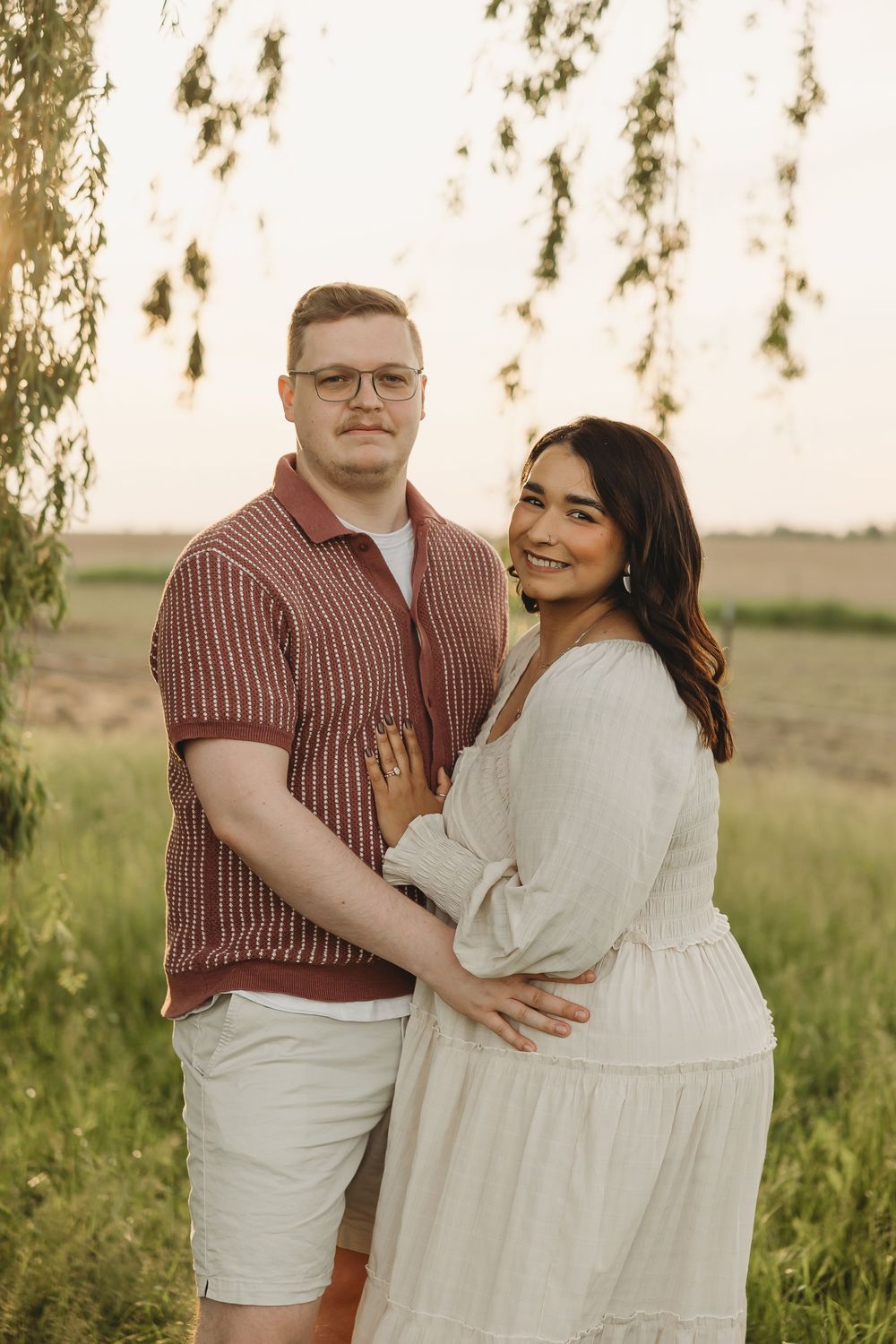 Couple embracing outdoors at golden hour, man in red shirt and woman in white dress, standing under willow tree.