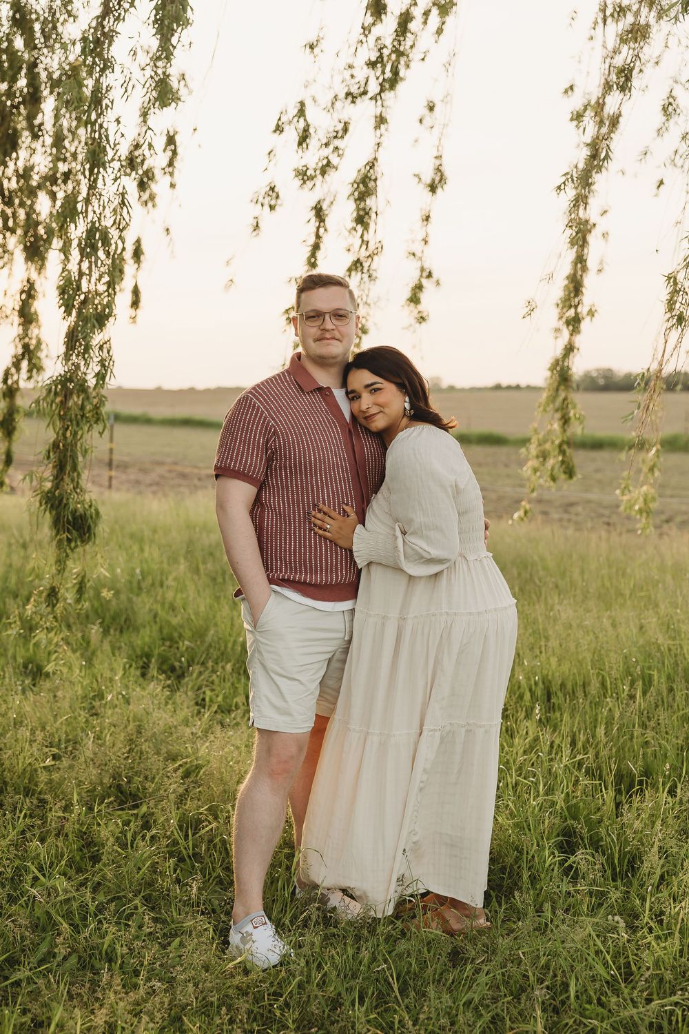 A couple embraces outdoors near a weeping willow tree in a green field at sunset, woman in white dress.