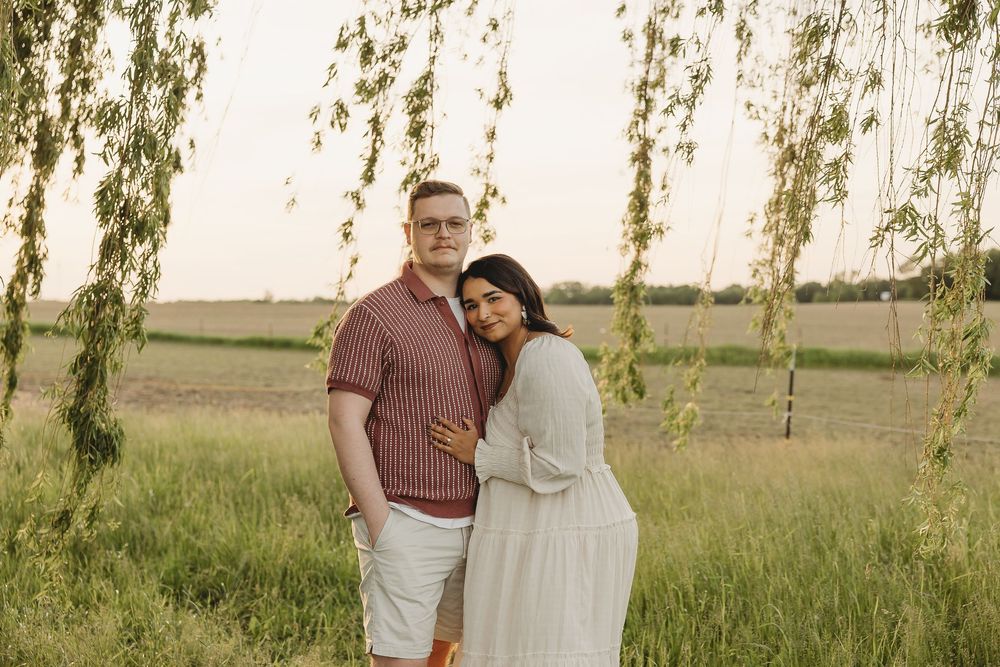 Couple embracing under weeping willow tree in green field at golden hour sunset.