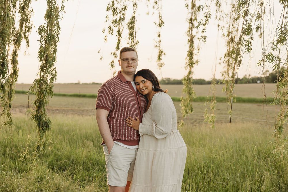 Couple embracing under weeping willow branches in a green field at golden hour, woman in white dress.