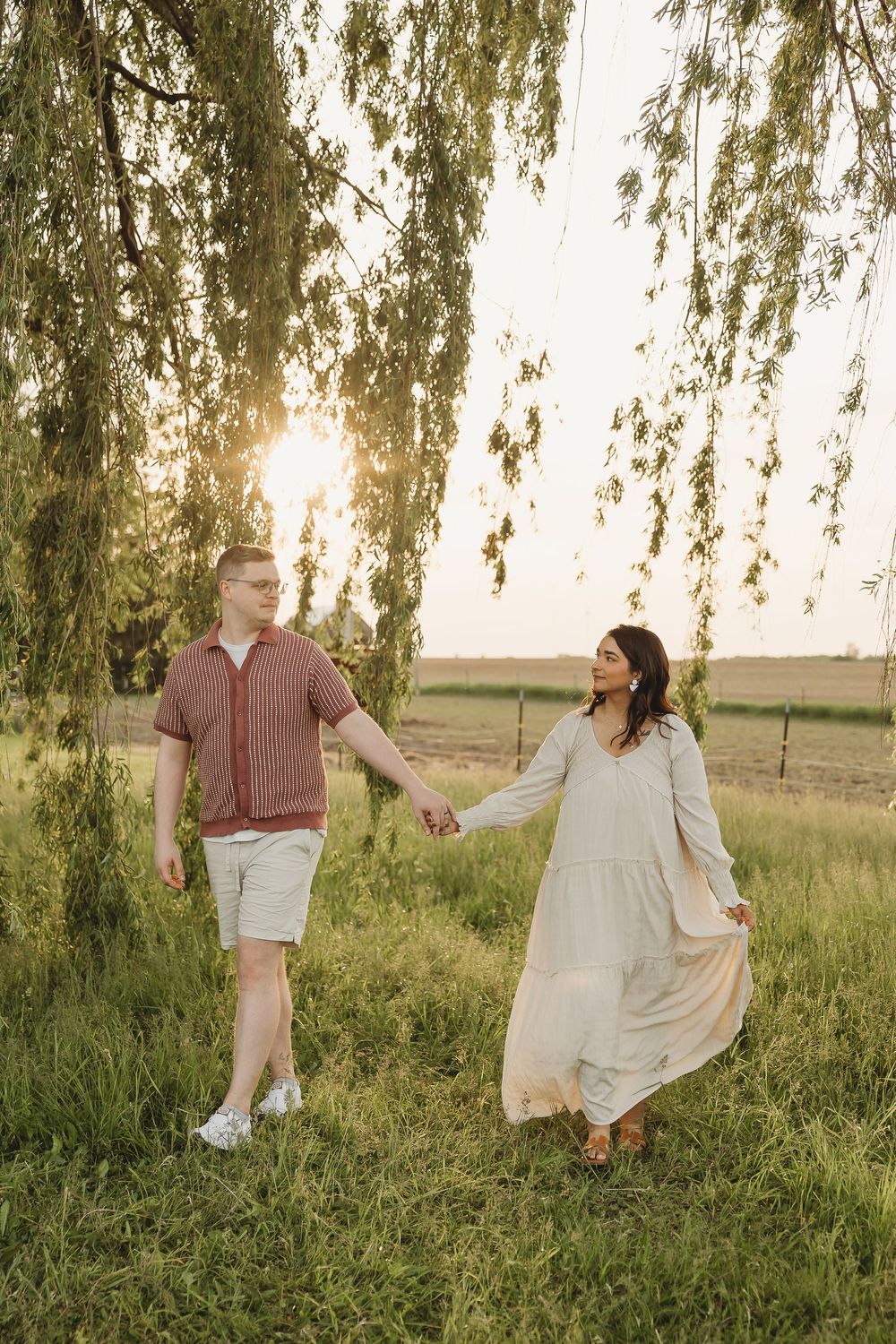 Couple holding hands walking under a weeping willow tree at sunset in a lush green field.