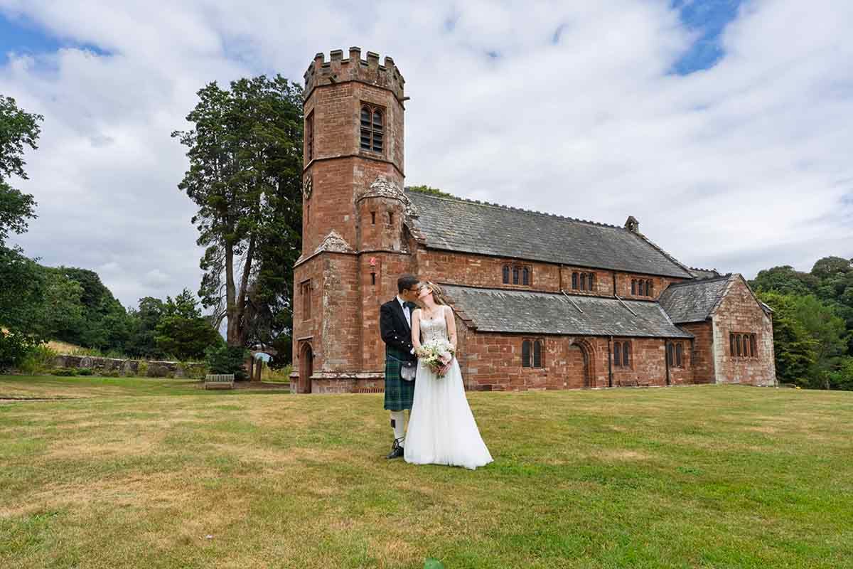 Bride and groom posing outside a historic red sandstone church with a tower on their wedding day.