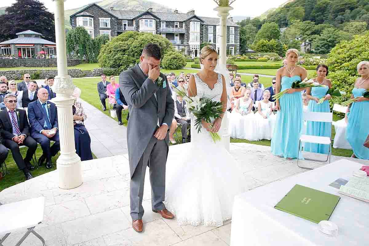 Emotional groom wiping tears as bride holds bouquet during outdoor wedding ceremony with guests seated on a sunny day.