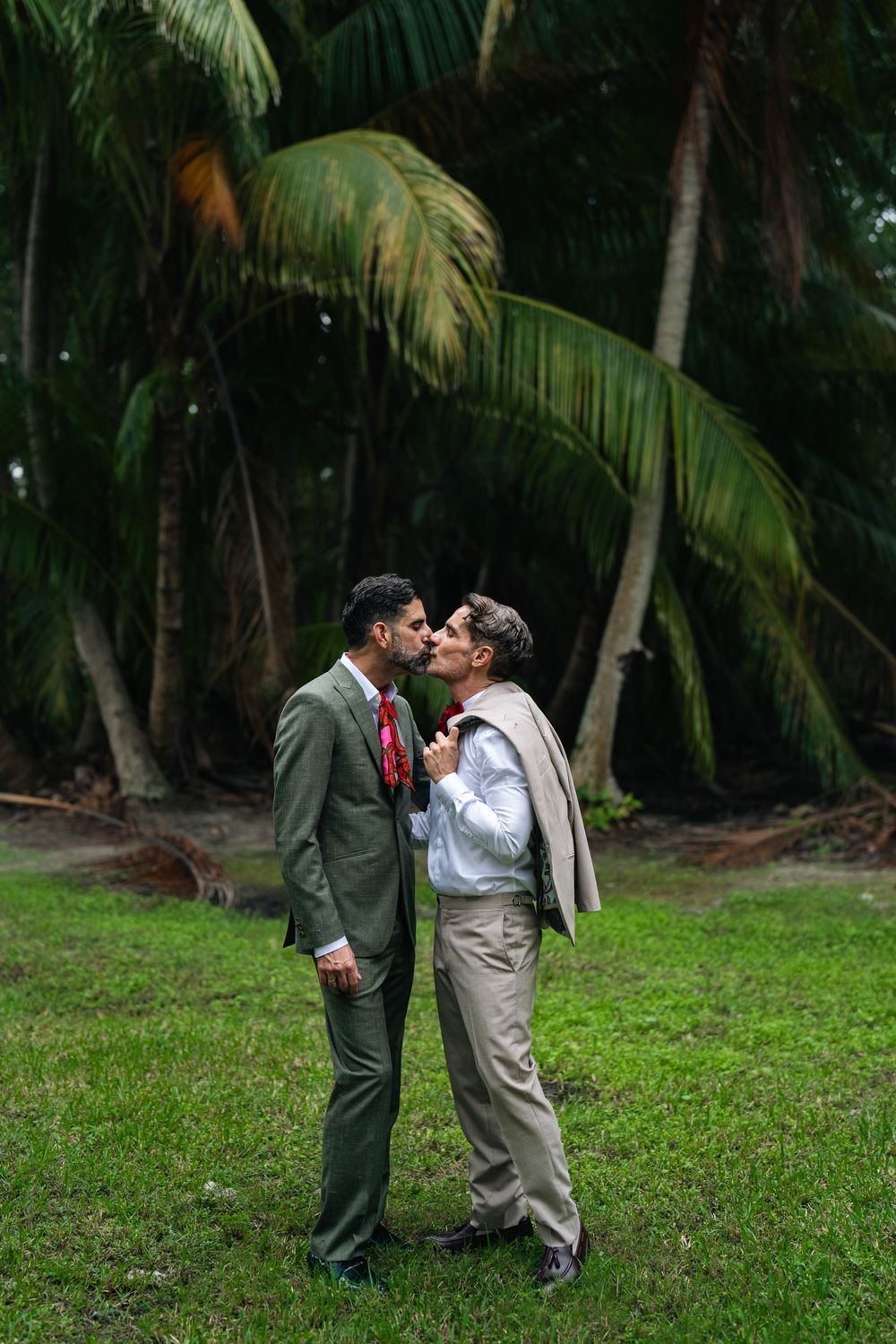 Two grooms sharing a kiss outdoors surrounded by lush tropical palm trees on green grass.