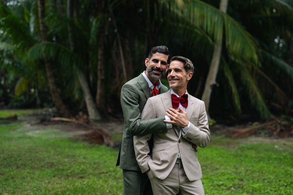 Two grooms in suits embrace outdoors among palm trees, smiling joyfully on their wedding day.