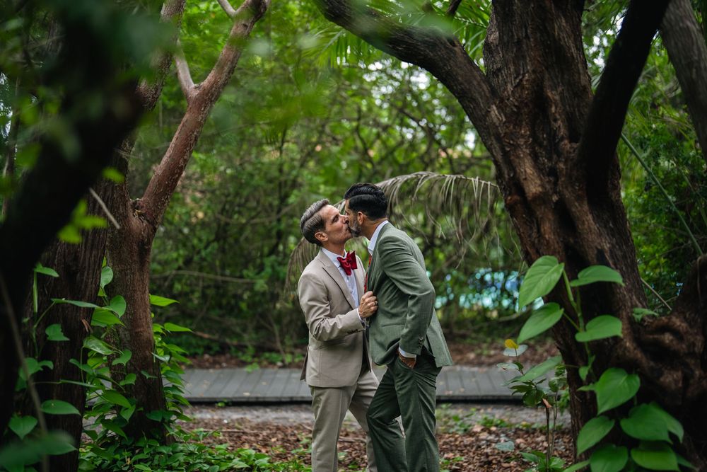 Two grooms in suits share a romantic kiss surrounded by lush green trees in an outdoor garden setting.