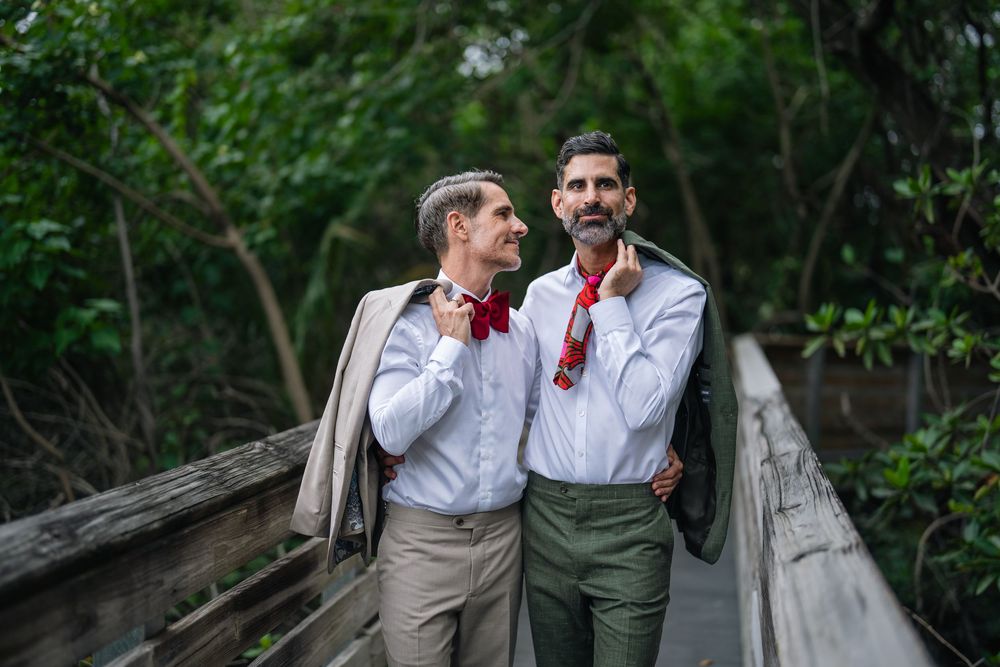 Two grooms in white shirts and red bow ties hold jackets on a wooden bridge surrounded by lush green trees.