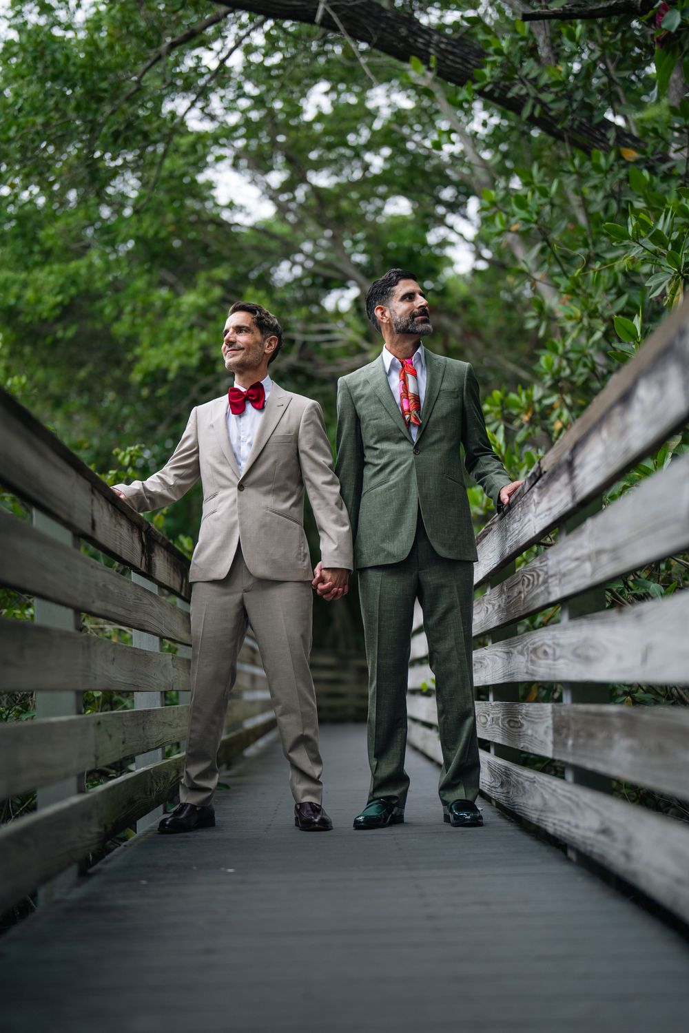 Two grooms in suits holding hands on a wooden bridge surrounded by lush greenery on their wedding day.
