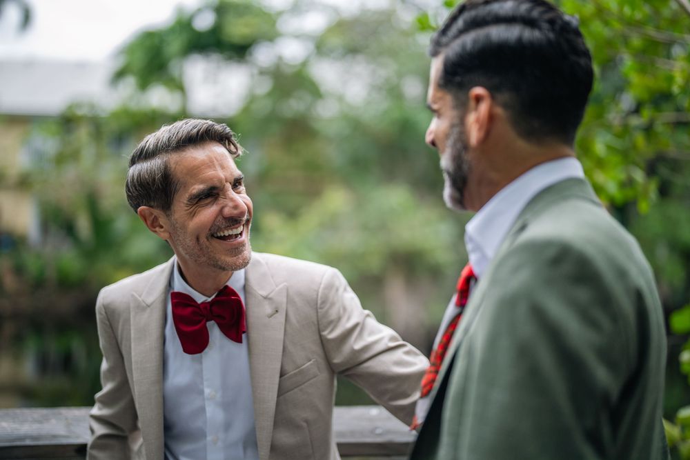 Two men in formal attire with red bow ties share a cheerful conversation outdoors.