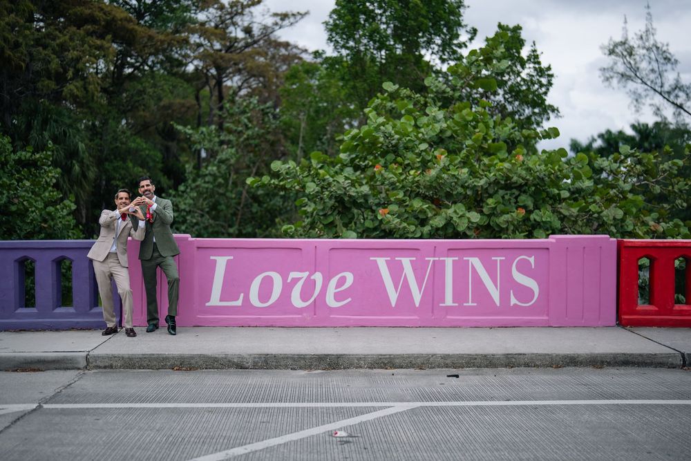 Two grooms pose together in front of a pink wall with Love Wins sign at an outdoor wedding venue.