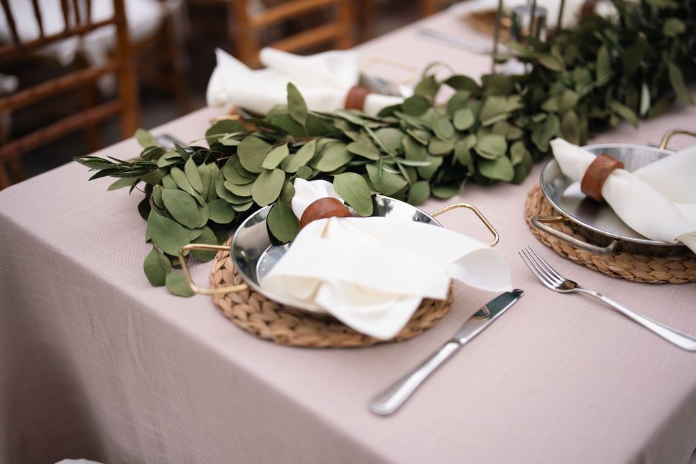 Elegant wedding table setting with wicker chargers, white napkins, greenery runner, and silver cutlery on a taupe linen.