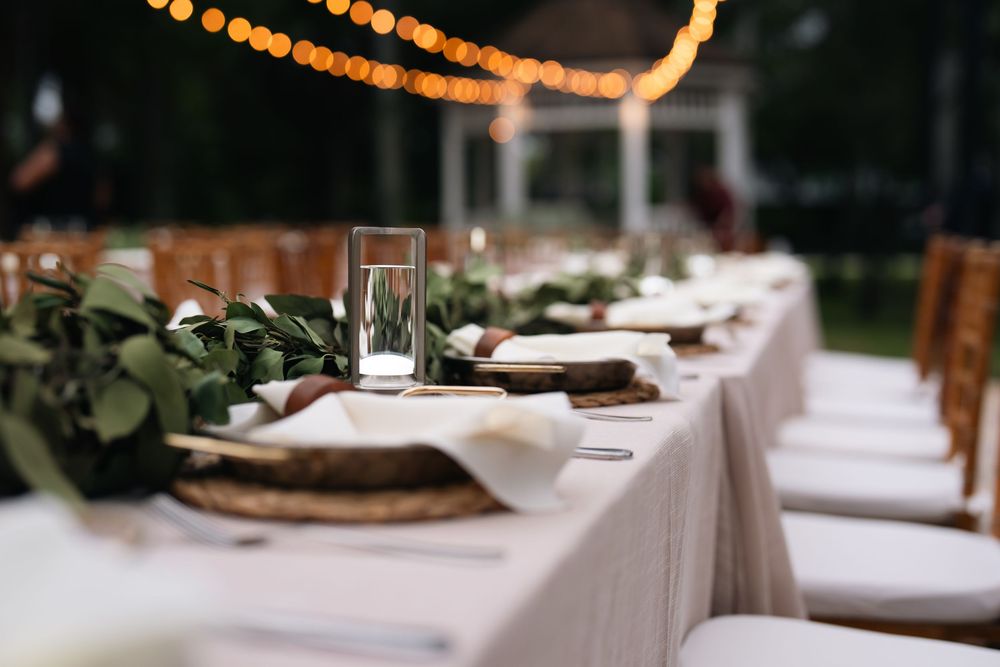 Elegant outdoor wedding reception table with greenery runner, white linens, and warm string lights at dusk.