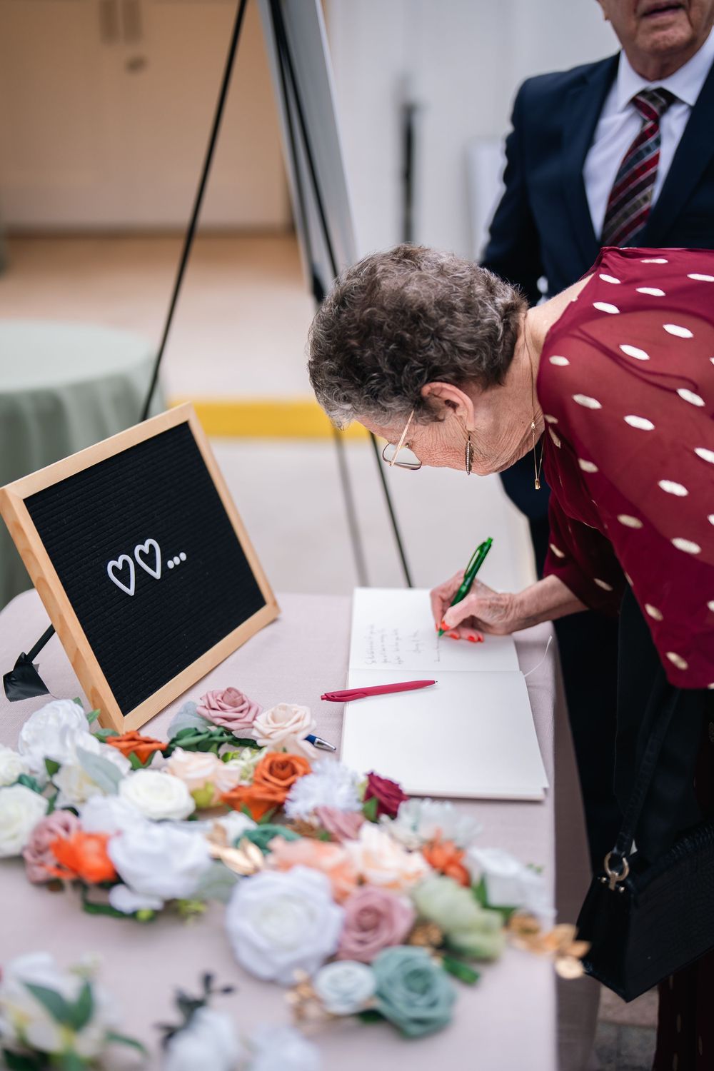 Elderly woman signing a wedding guest book at a decorated table with floral arrangements and a heart chalkboard sign.