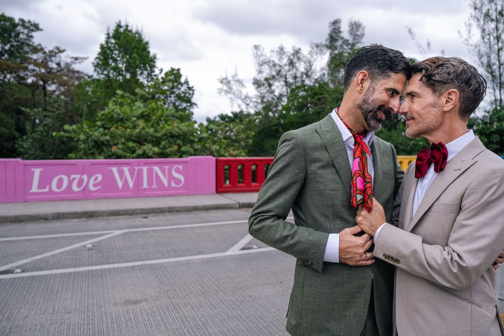 Two grooms in suits embrace in front of a pink Love Wins sign, celebrating their wedding day outdoors.
