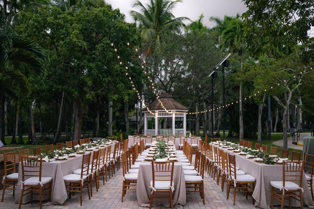Elegant outdoor wedding reception with string lights, long tables, chiavari chairs, and lush tropical trees in background.