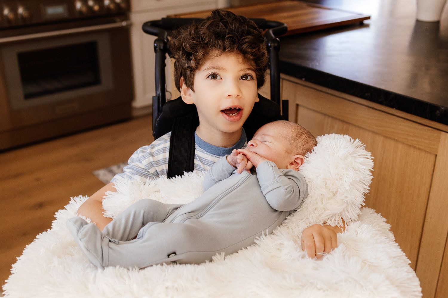 Young boy in wheelchair proudly holding sleeping newborn baby brother on white fluffy blanket in kitchen during their in-home photo session.