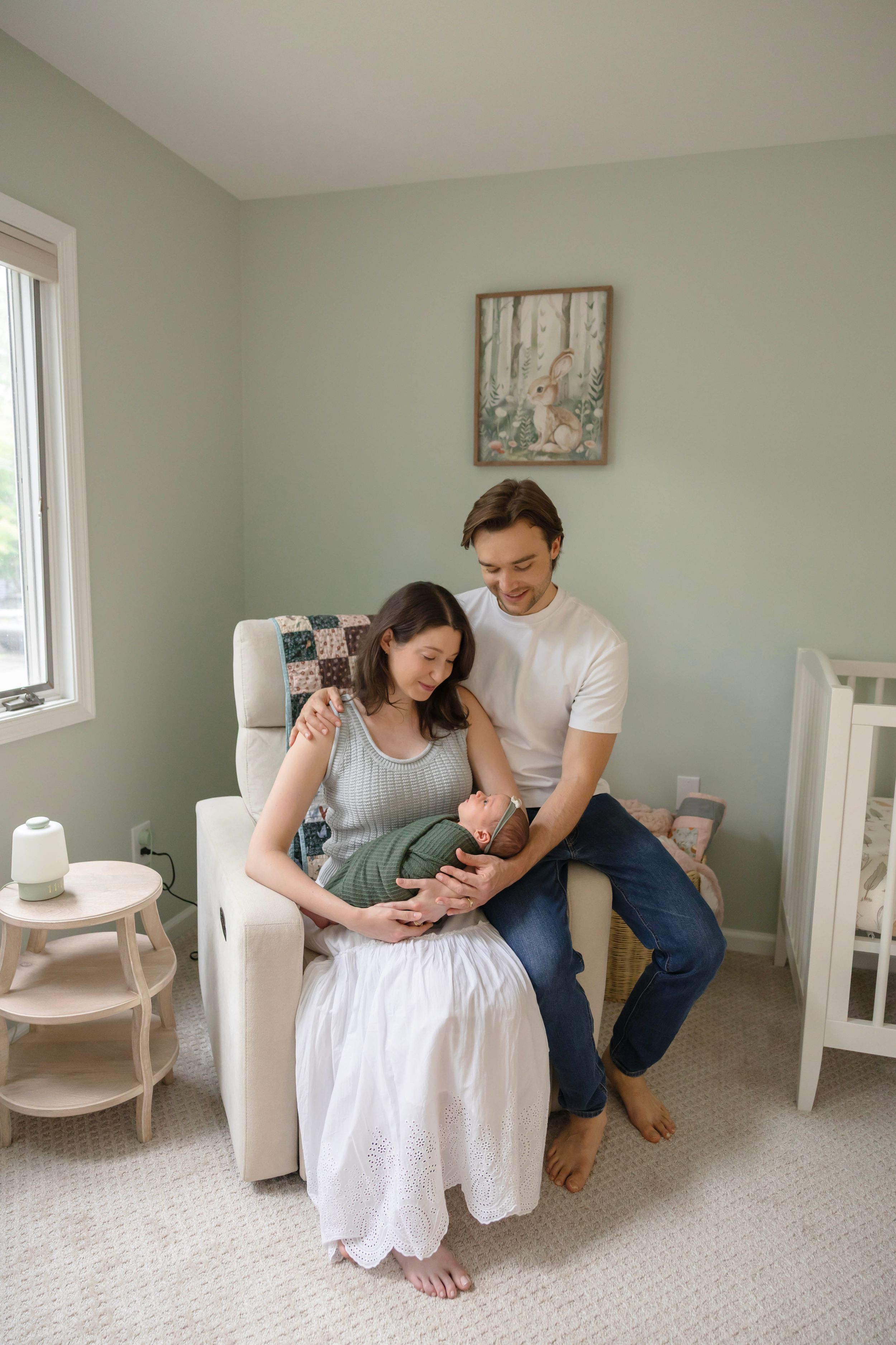 New parents sitting in nursery chair holding their swaddled newborn baby in a green wrap.