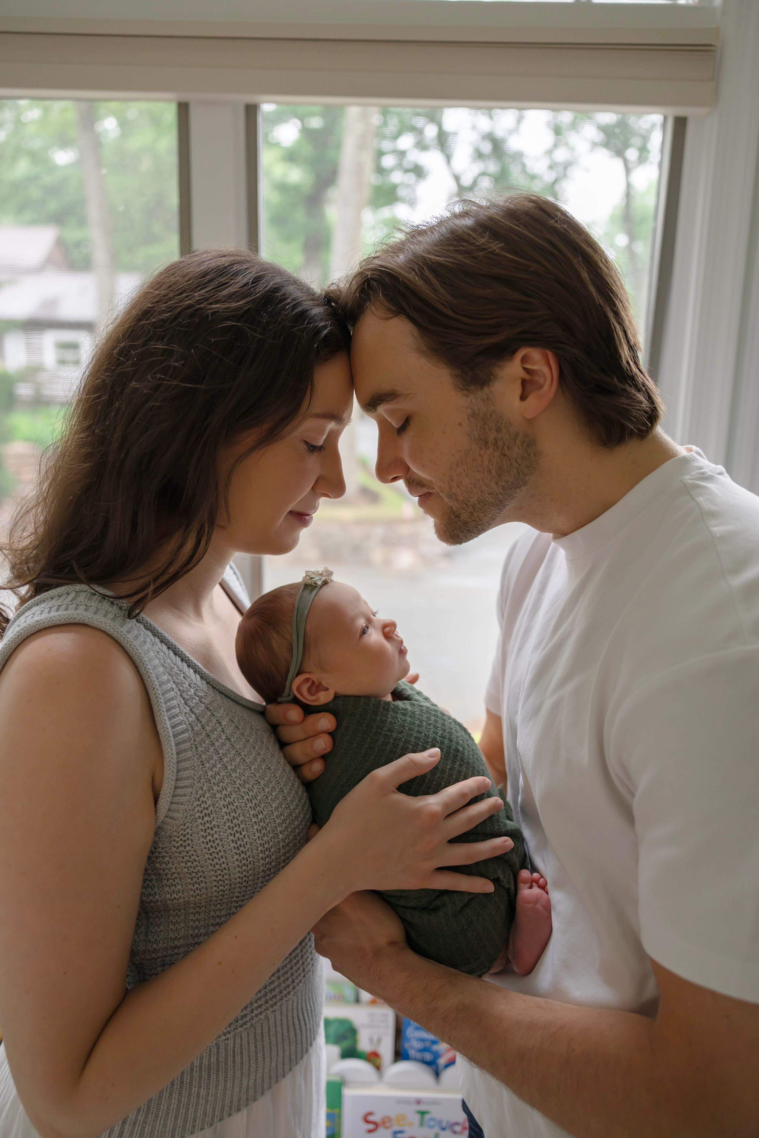 New parents tenderly touch foreheads while cradling their swaddled newborn baby in a green wrap near a window.