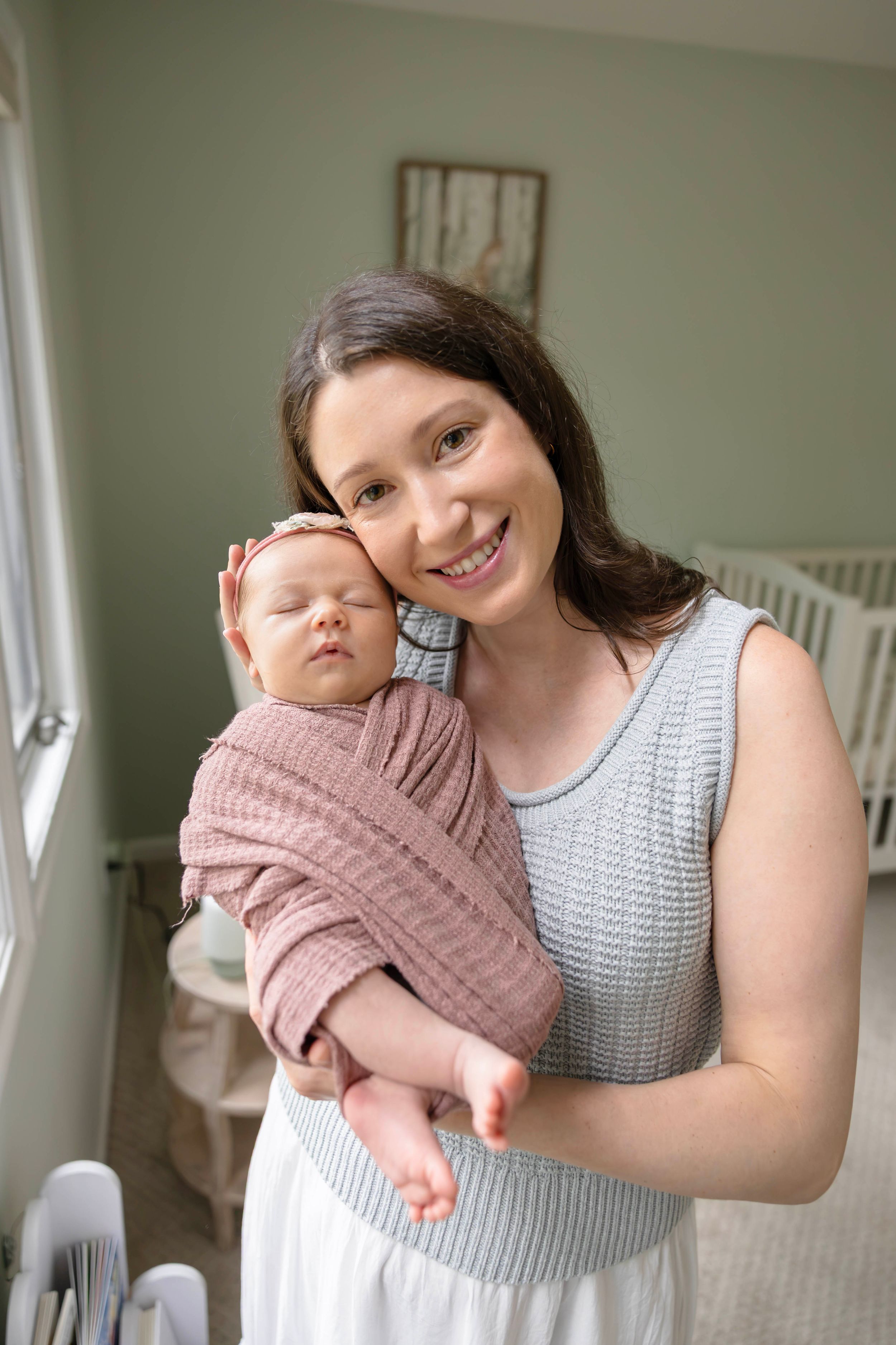 Smiling young mother holding sleeping newborn baby wrapped in pink swaddle blanket in nursery room.