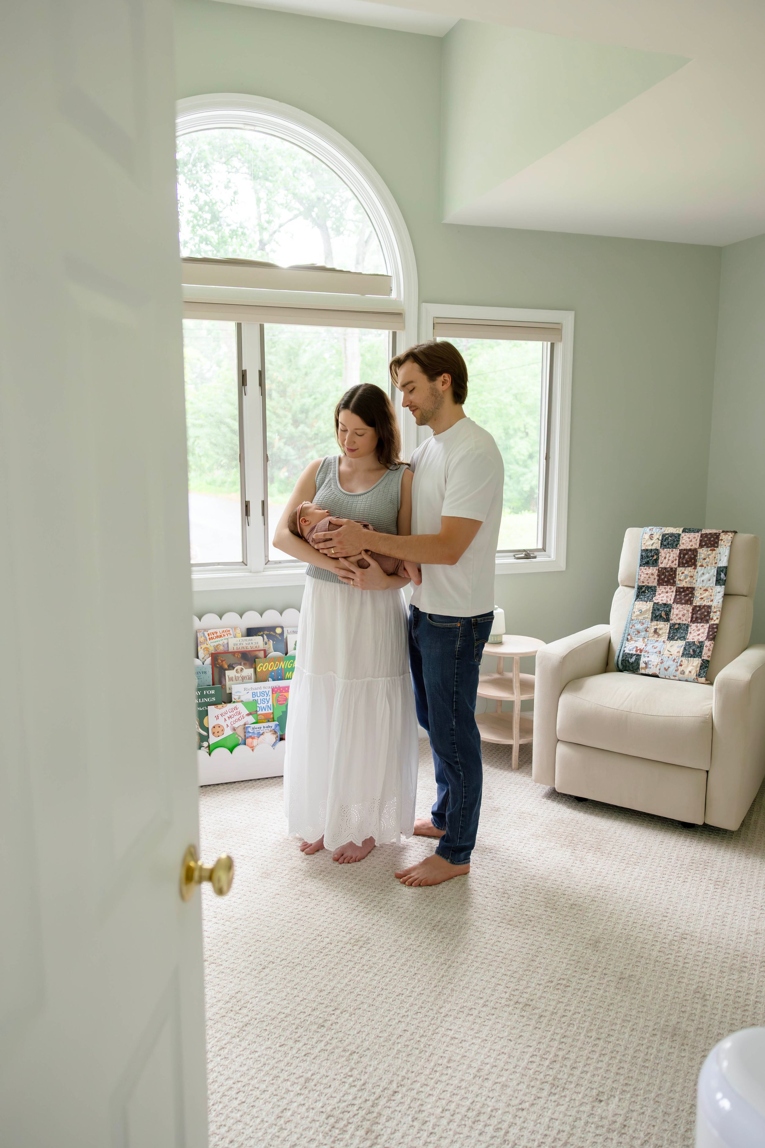 New parents stand together in a nursery, lovingly gazing at their newborn baby near a bright arched window.