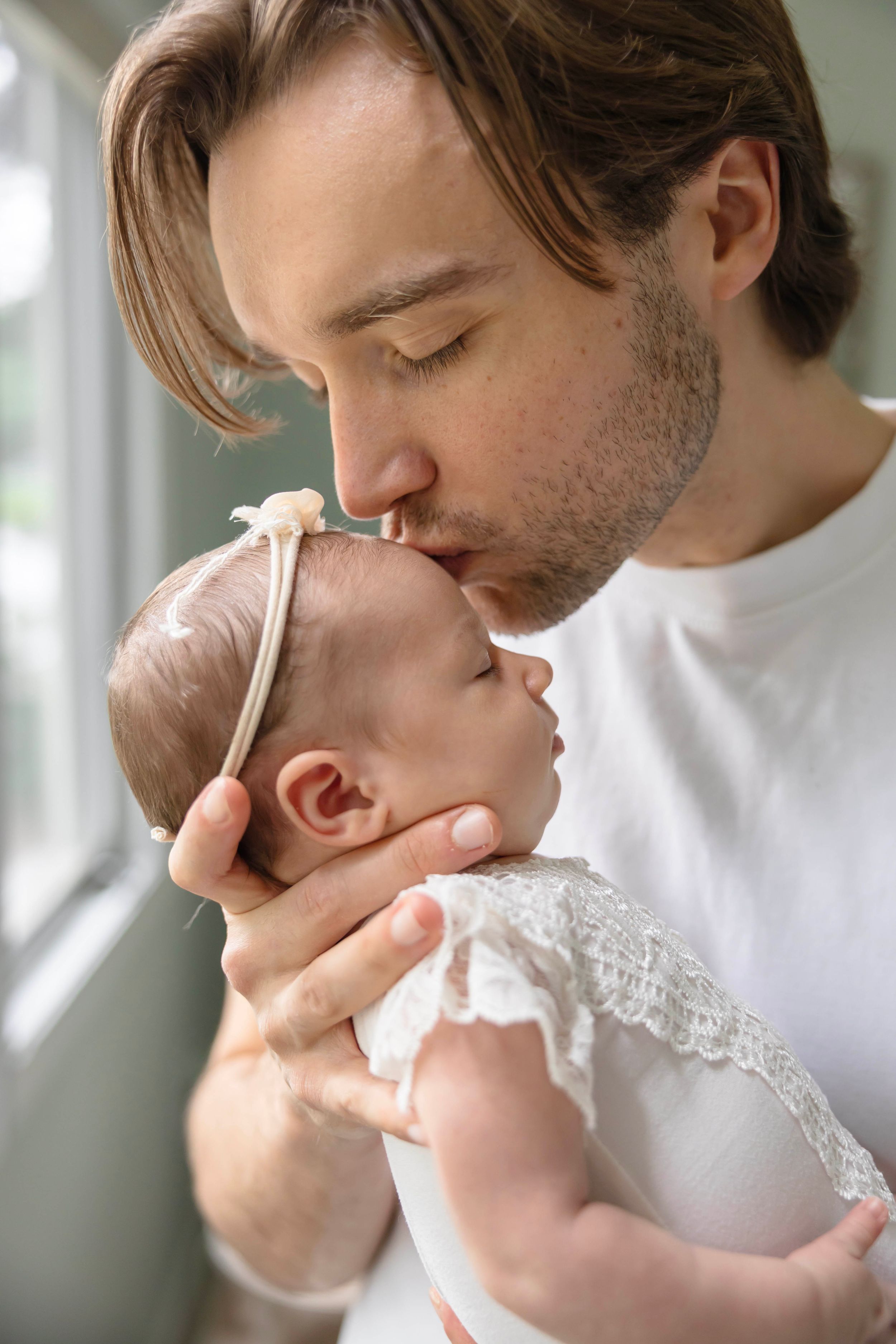 Father tenderly kissing his newborn baby girl dressed in white lace, holding her gently near a window.
