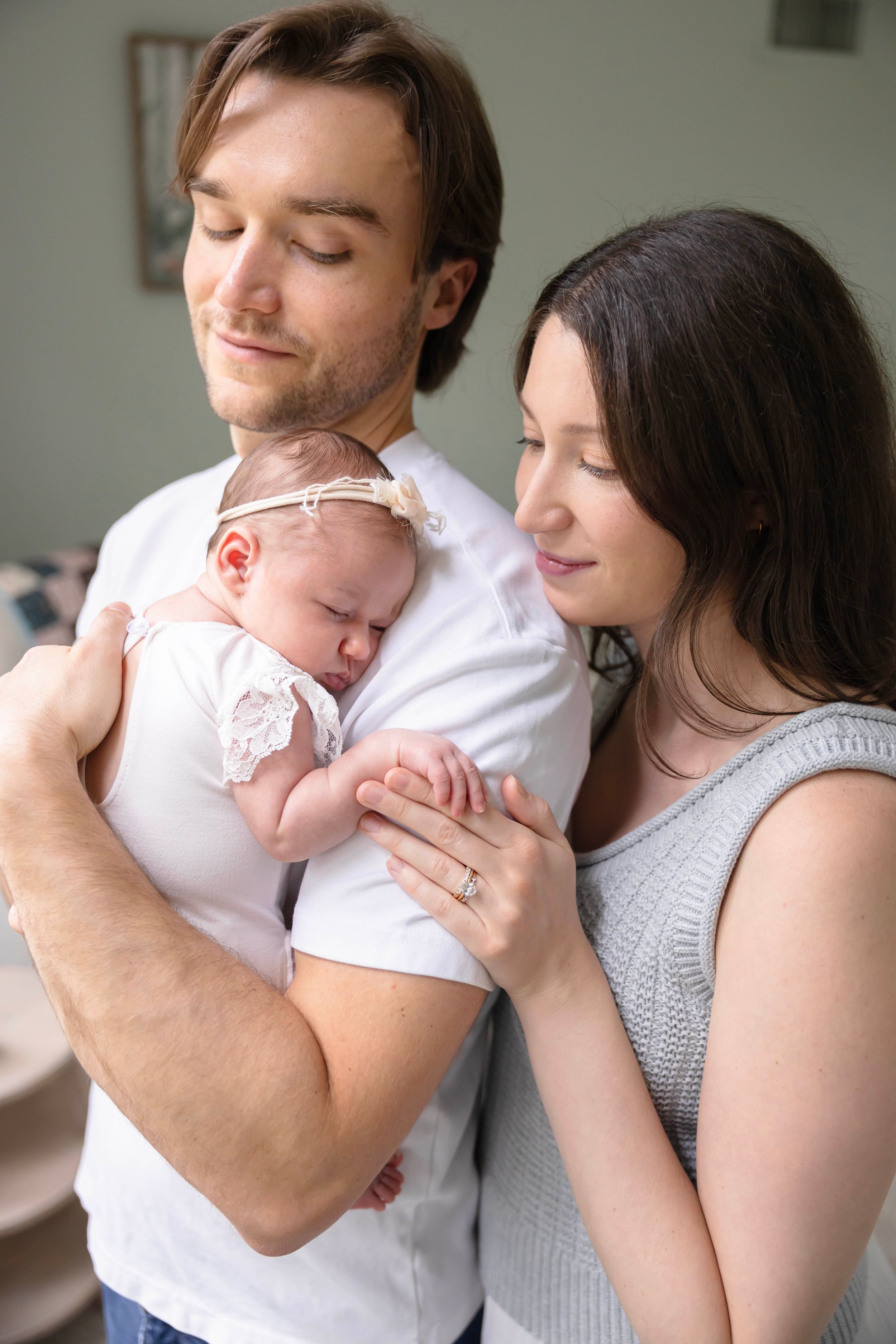Young parents tenderly holding their sleeping newborn baby girl wearing a white dress and floral headband indoors.